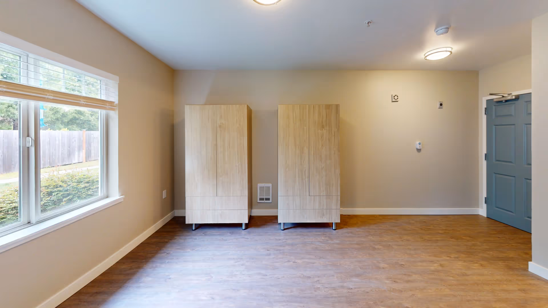 Empty senior living apartment room with two wooden wardrobes against a beige wall, a large window on the left and a blue door on the right.