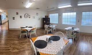 A dining room with several small tables covered with lace and patterned tablecloths, each set with placemats and napkins. The room has wooden flooring, large frosted windows letting in natural light, and wall decorations including framed pictures and a decorative sign that says 'Cafe'.