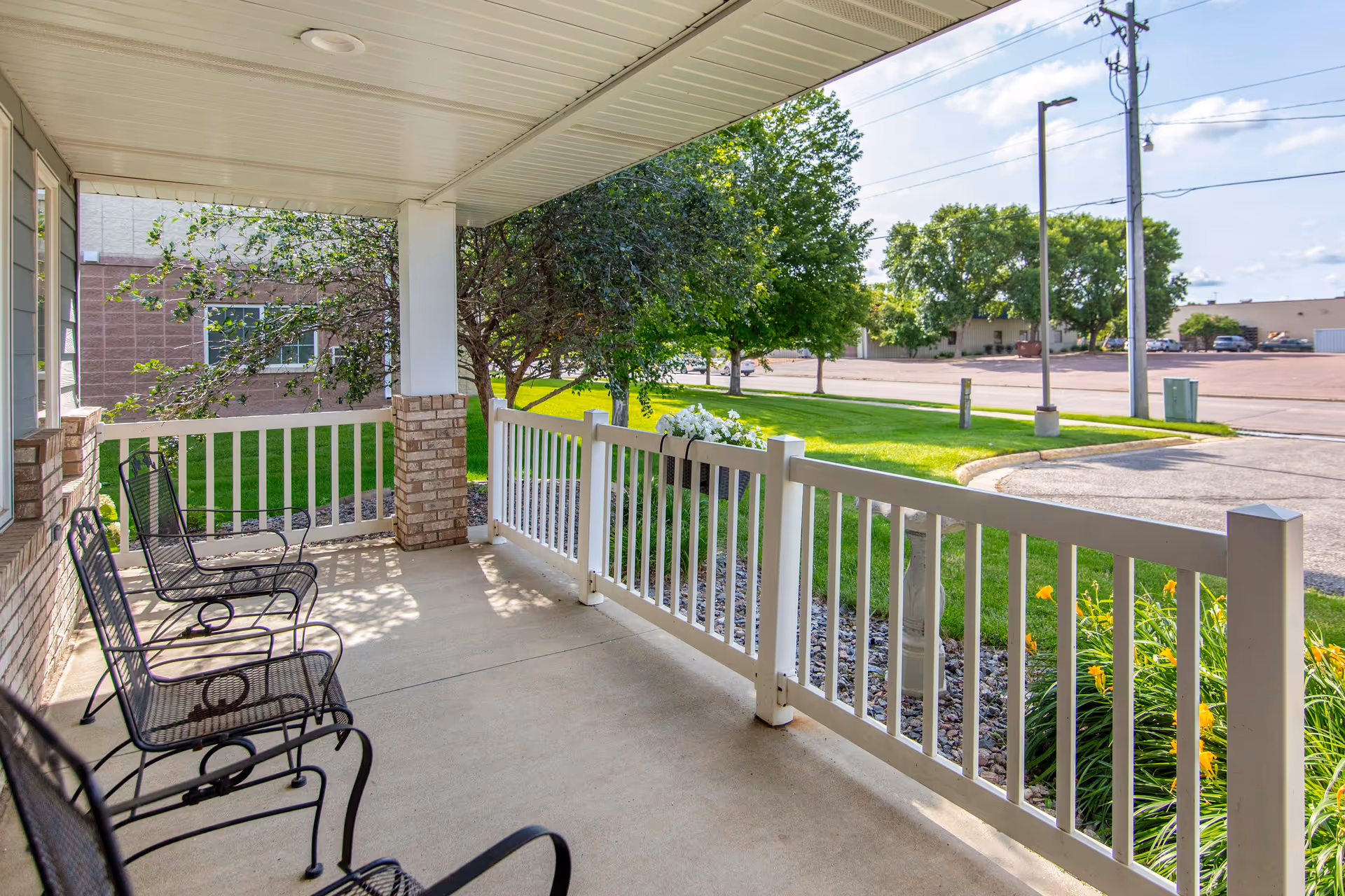 Covered front porch with metal chairs and a white railing overlooking a grassy lawn and street.