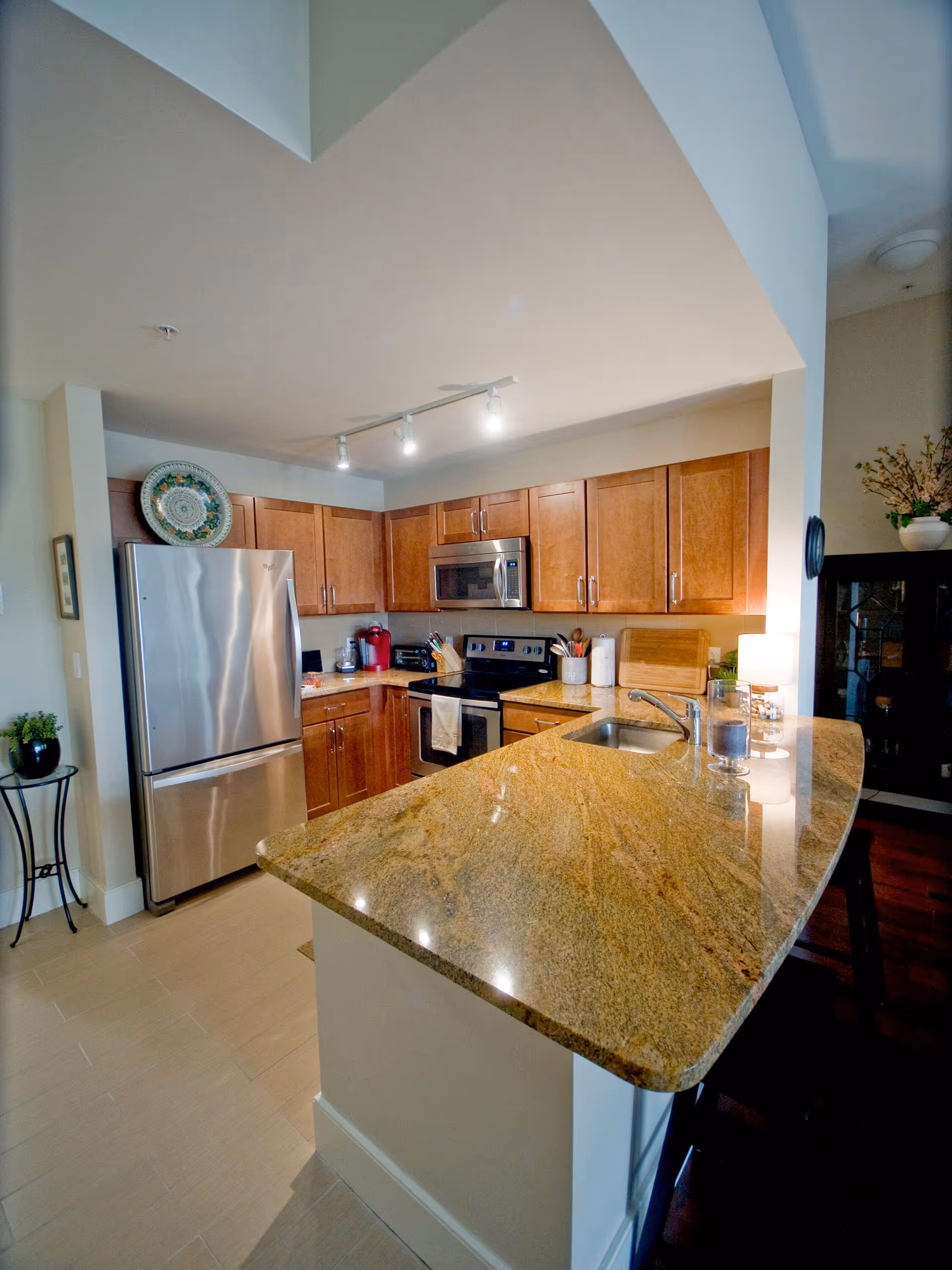 Modern kitchen with wooden cabinets, stainless steel refrigerator, microwave, and stove. The kitchen features a large granite countertop with a built-in sink and a few decorative items including a lamp, a glass candle holder, and a small plant on a side table.
