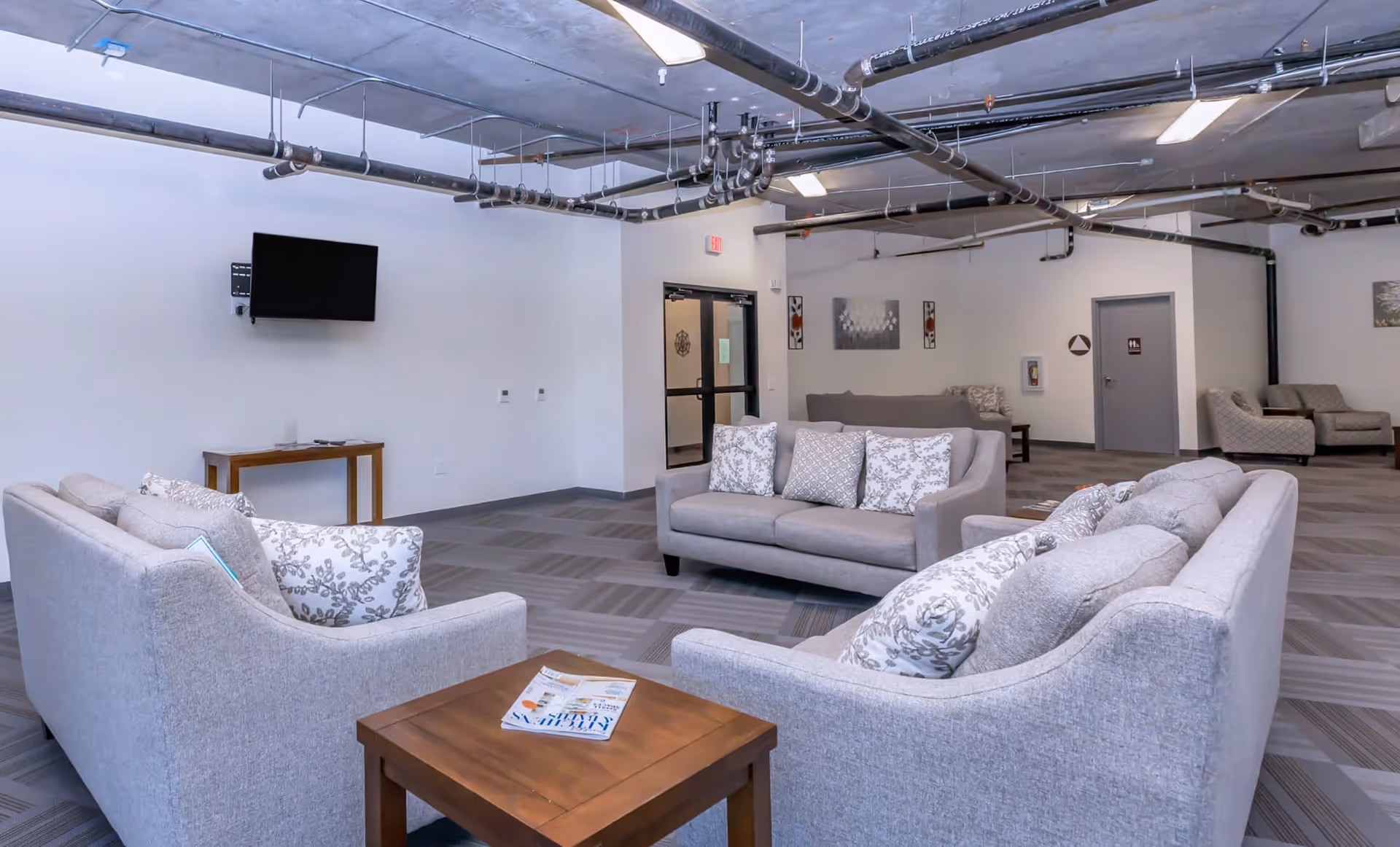 A spacious lounge area with multiple light gray sofas adorned with patterned cushions arranged around wooden tables. The room has a modern industrial ceiling with exposed pipes and fluorescent lighting. A flat-screen TV is mounted on a white wall above a small wooden console table. In the background, there are additional seating areas, a door, and some wall decorations.