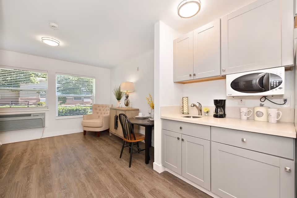 Bright studio-style room with a kitchenette (sink, microwave, coffee maker) on the right and a small seating area with a table and armchair by the windows on the left.
