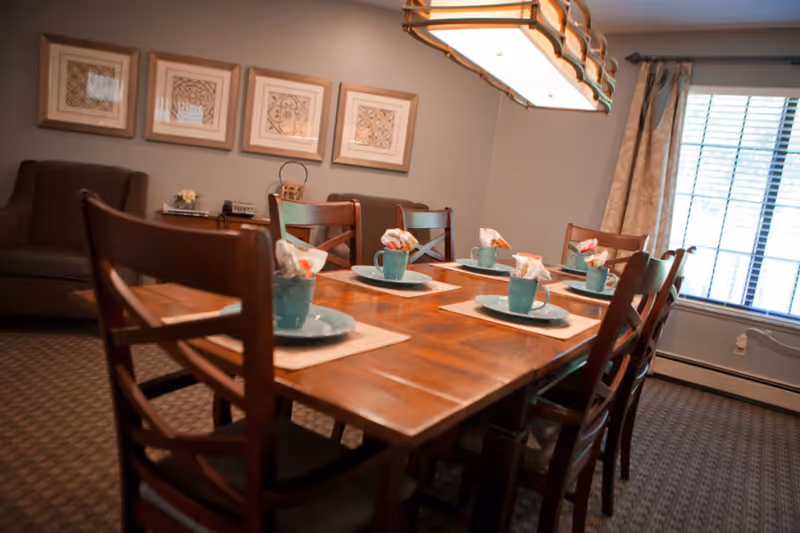 A wooden dining table set for six with turquoise cups and napkins in a cozy dining room with a hanging light and window.