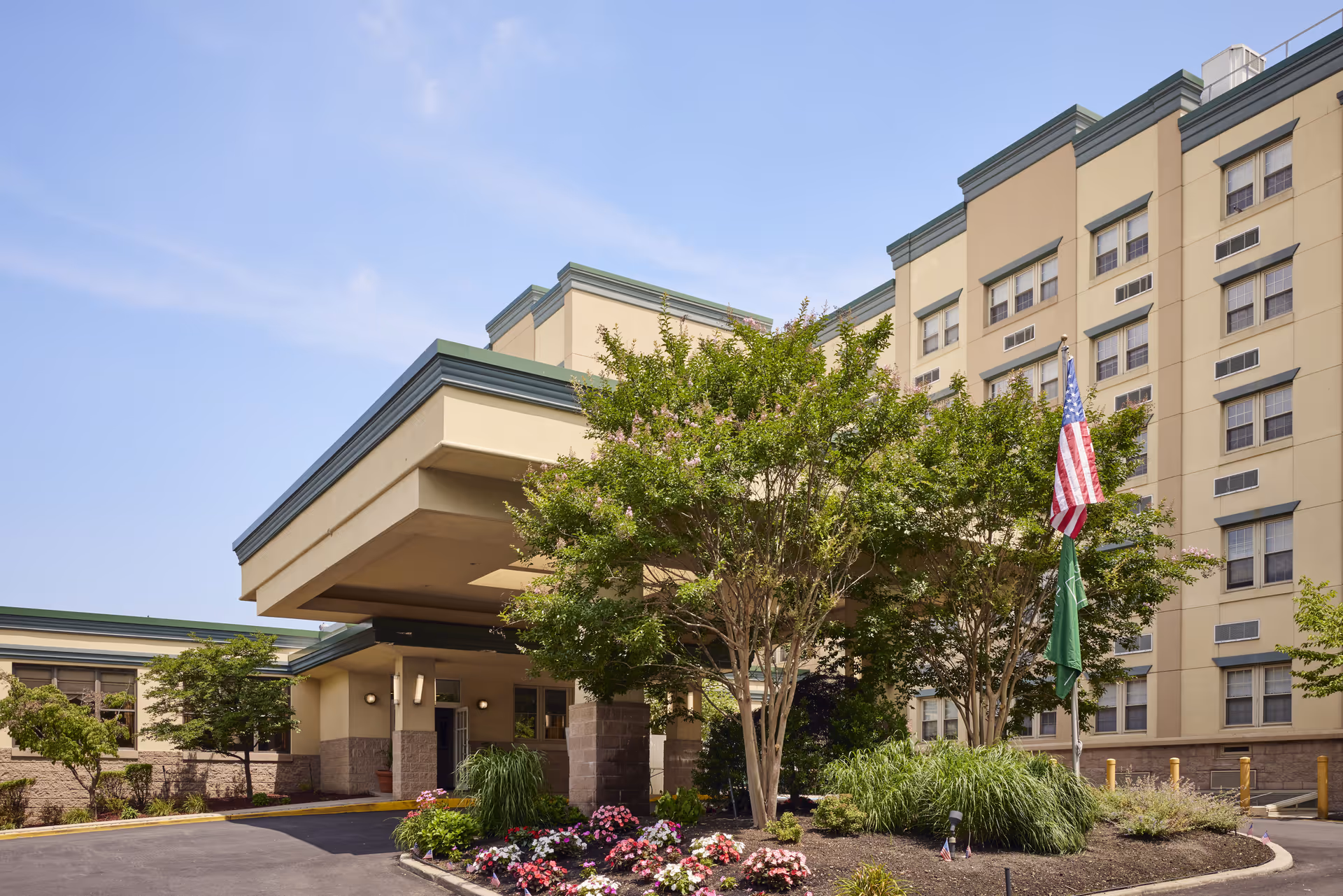 Exterior view of Brandywine The Savoy by Monarch senior living facility showing the entrance with a covered drop-off area, landscaped garden with flowering plants and trees, and two flagpoles displaying the American flag and another green flag.