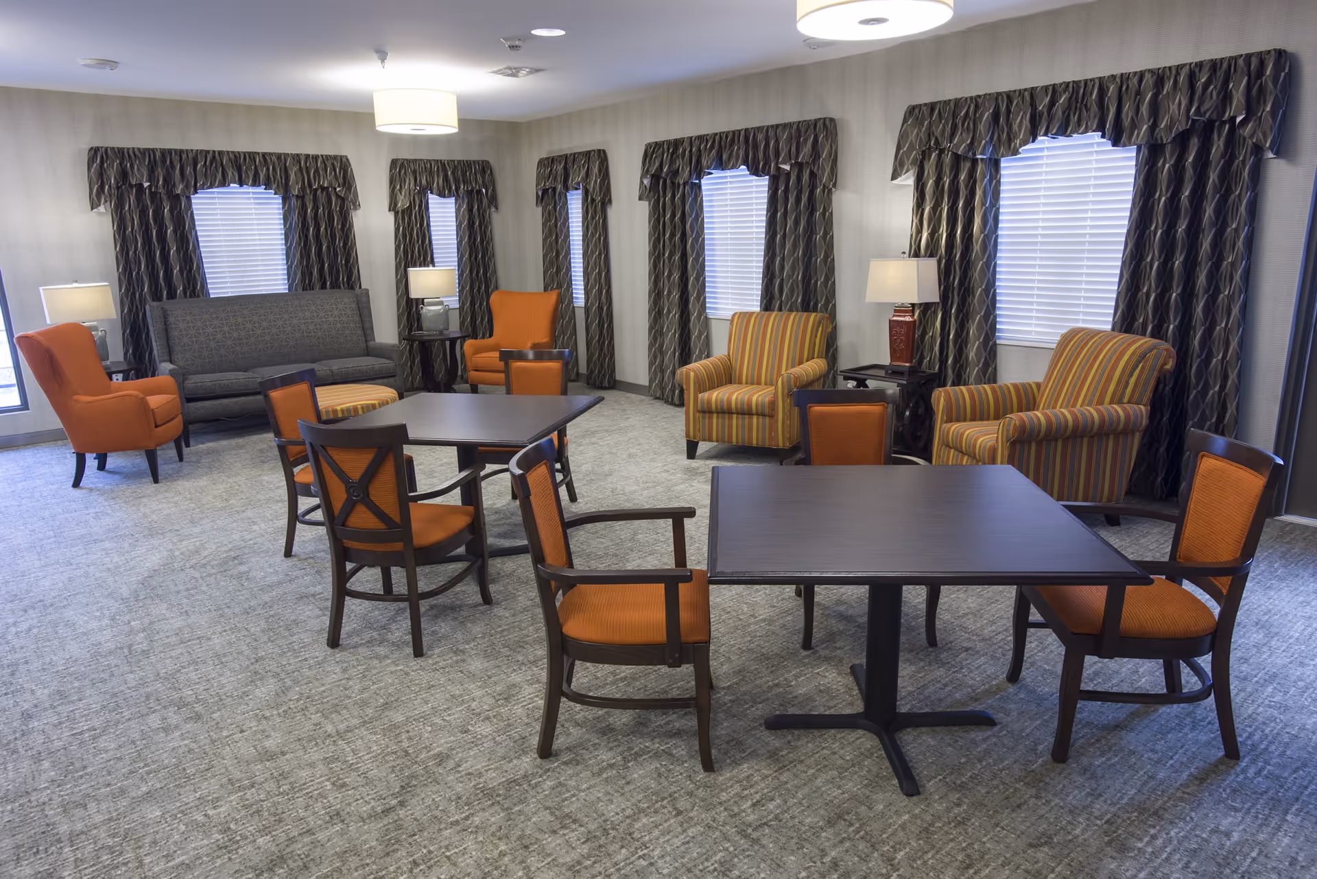 A spacious common room with multiple seating options including orange cushioned chairs, striped armchairs, and a gray sofa. There are two square tables with orange cushioned chairs around them. The room has several windows with dark patterned curtains and blinds, and is softly lit by ceiling lights and table lamps.