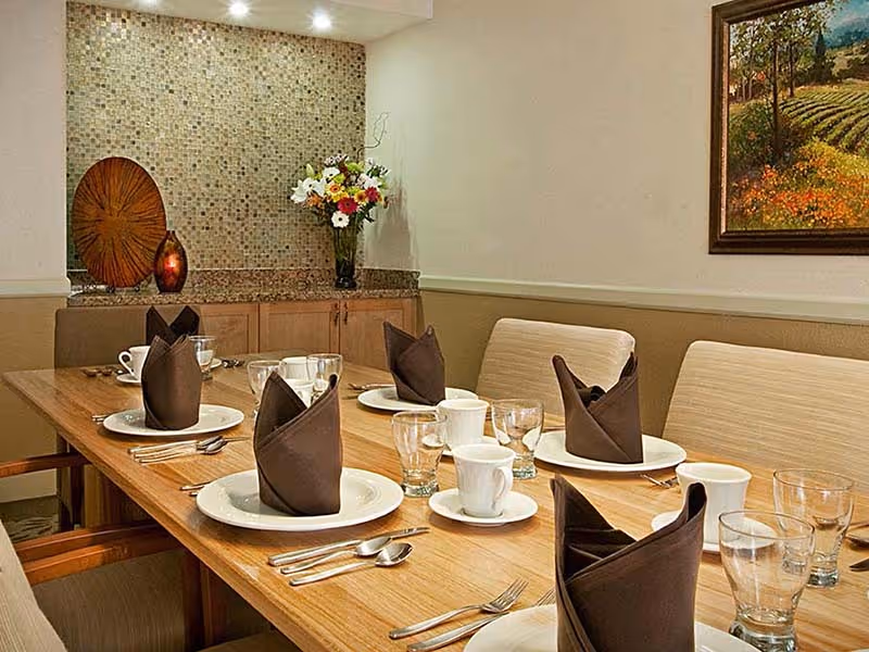 A dining room table set for six with white plates, cups, glasses, and brown folded napkins. The table is wooden with beige cushioned chairs around it. In the background, there is a mosaic tiled wall with a decorative wooden piece, a vase with colorful flowers, and a framed painting on the adjacent wall.