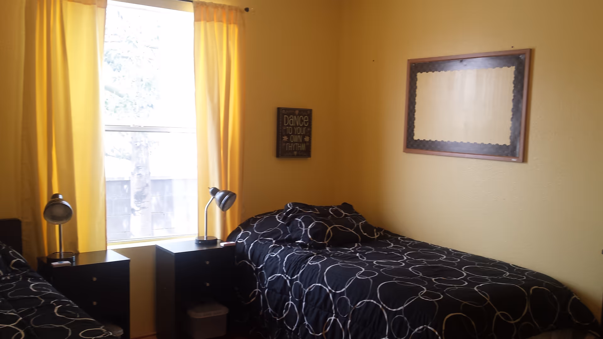 A bedroom with two single beds covered in black bedspreads with white circular patterns. Between the beds are two black nightstands, each with a silver desk lamp. A large window with yellow curtains lets in natural light. The walls are painted yellow and have two framed decorations, one with text and one blank.