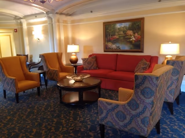 A cozy living room area with a red sofa, four patterned armchairs, a round wooden coffee table with a decorative bowl, two table lamps on side tables, and a framed landscape painting on the wall. The room has warm lighting and a patterned carpet.