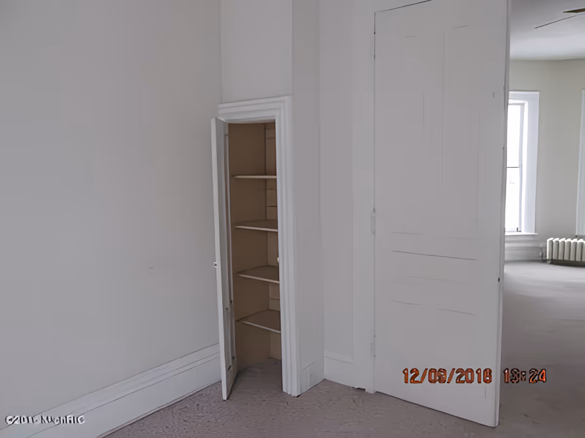 An empty interior room with white walls and carpeted floor. There is a small open closet with shelves in the corner and a partially open door leading to another room with windows and a radiator.