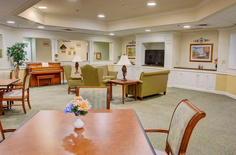 A bright senior living common room with tables and chairs in the foreground, sofas and armchairs around a TV, and a piano in the background.
