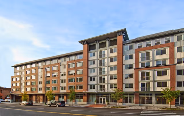 Exterior view of a modern multi-story residential building with a combination of brick and light-colored siding. The building has many windows and small balconies, with trees and parked cars along the street in front.