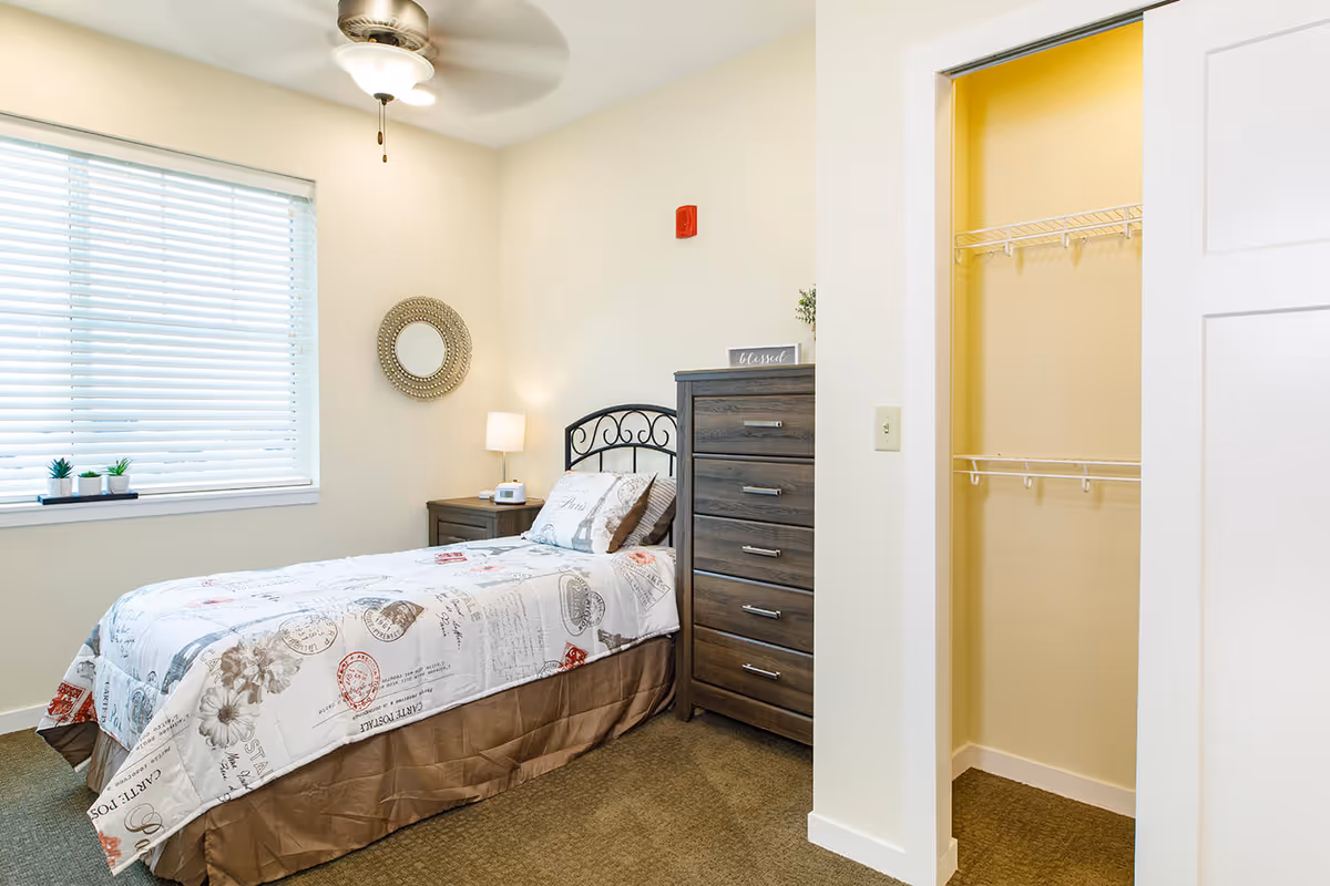 Furnished bedroom with a single bed, nightstand and lamp, tall dresser, ceiling fan, window with blinds, and an open closet.