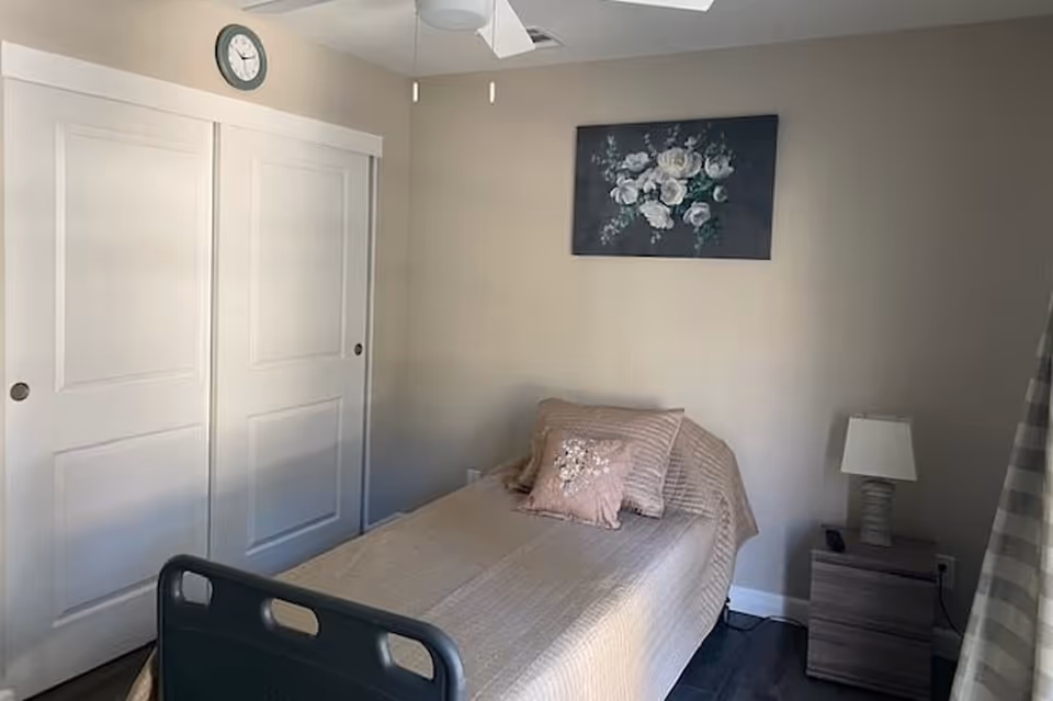 A small bedroom with a single bed covered in beige bedding and two decorative pillows. There is a nightstand with a lamp and a remote control next to the bed. On the wall above the bed is a painting of white flowers. A clock is mounted on the wall above white sliding closet doors. The room has neutral-colored walls and a ceiling fan.