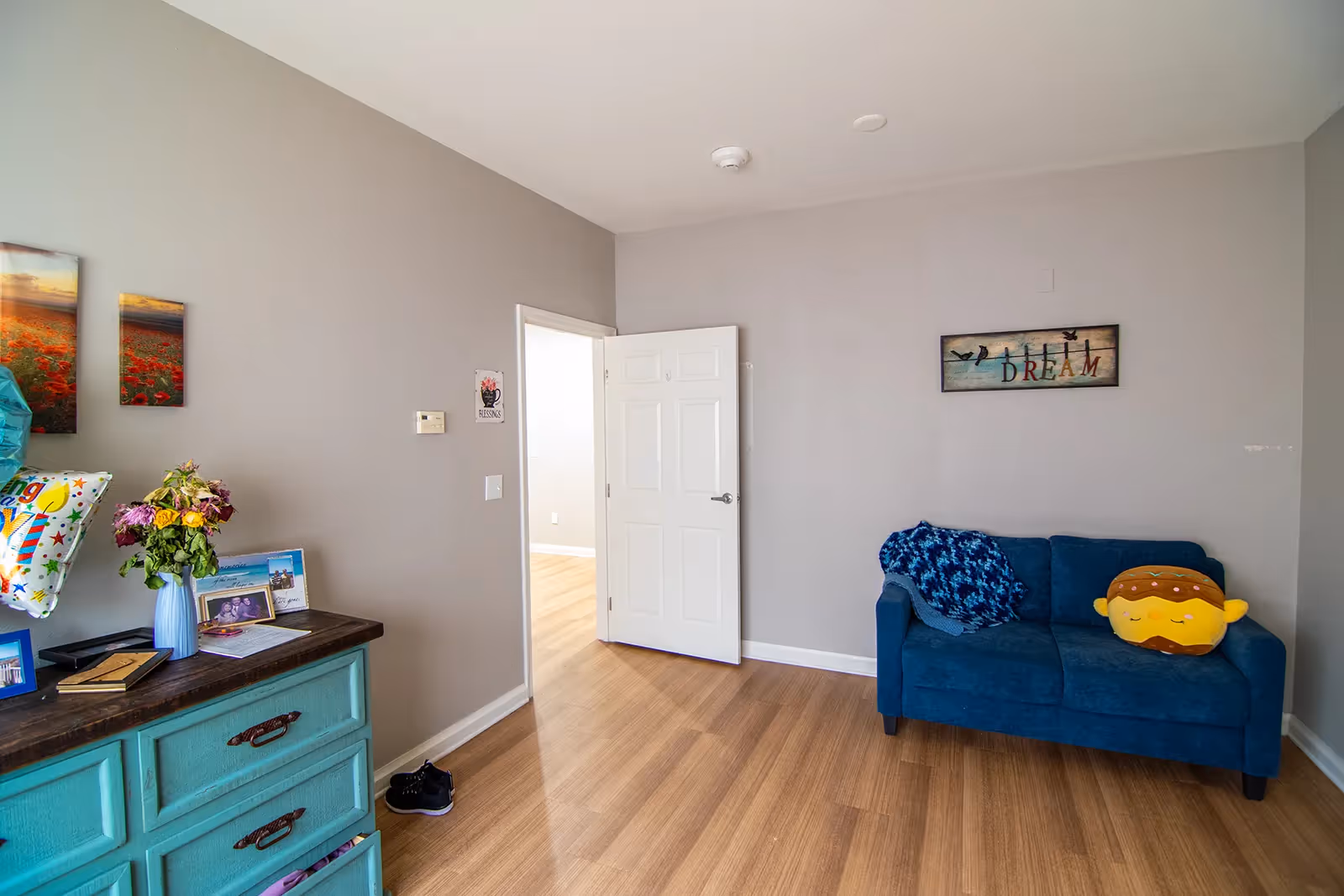 A small living room with light gray walls and wood flooring. There is a blue loveseat with a blue patterned throw and a plush hamburger pillow. On the wall above the loveseat is a framed artwork with birds and the word 'DREAM'. To the left, there is a turquoise dresser with a vase of flowers, framed photos, and balloons. An open white door leads to another room with similar flooring.