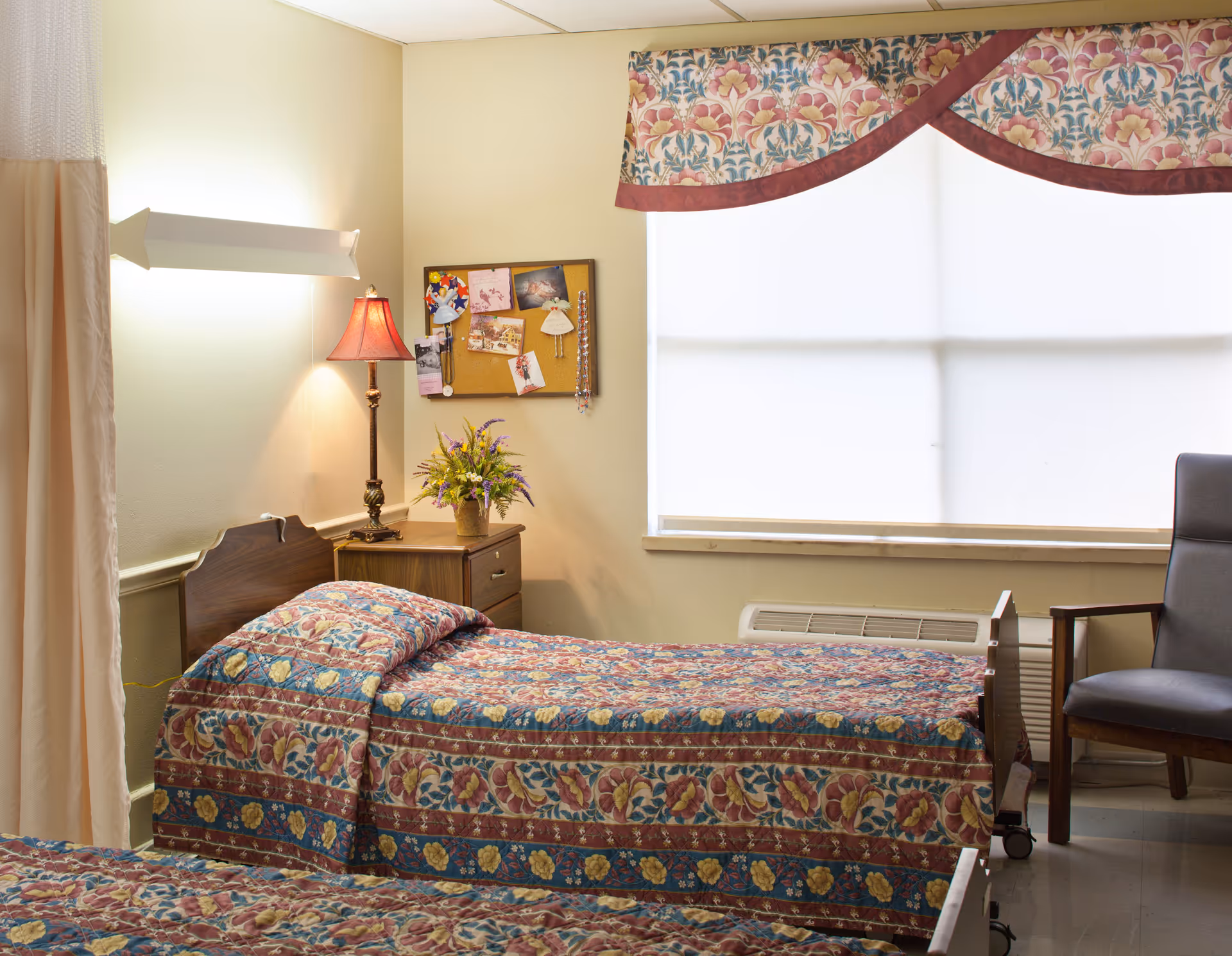A senior living facility bedroom with two single beds covered in floral patterned bedspreads. There is a wooden nightstand between the beds with a decorative lamp and a vase of flowers. A bulletin board with photos and cards is mounted on the wall above the nightstand. A large window with a floral valance lets in natural light, and a gray armchair is positioned near the window.