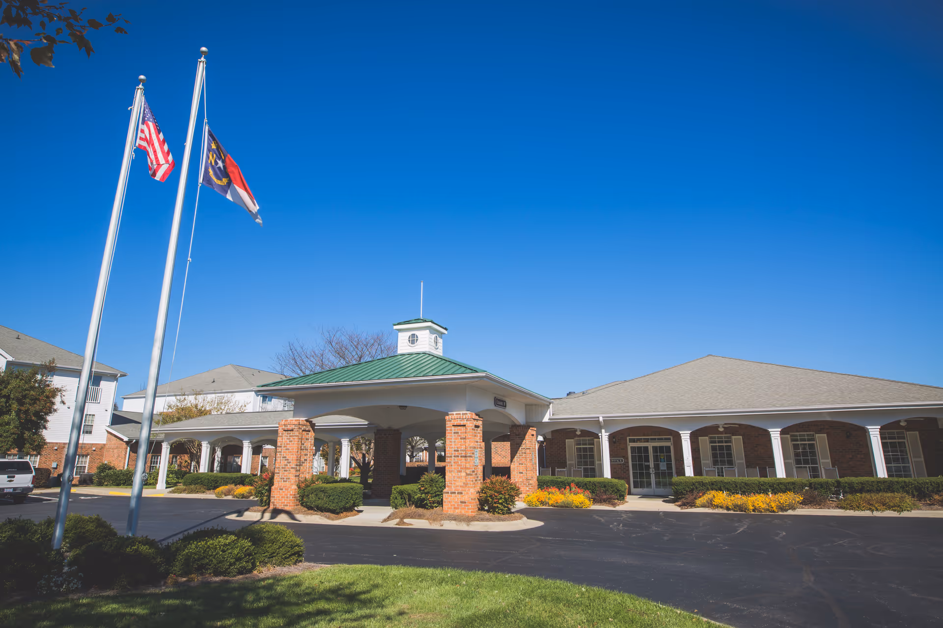 Front entrance of RidgeCrest with flagpoles, a covered porte-cochère, and landscaped driveway under a clear blue sky.