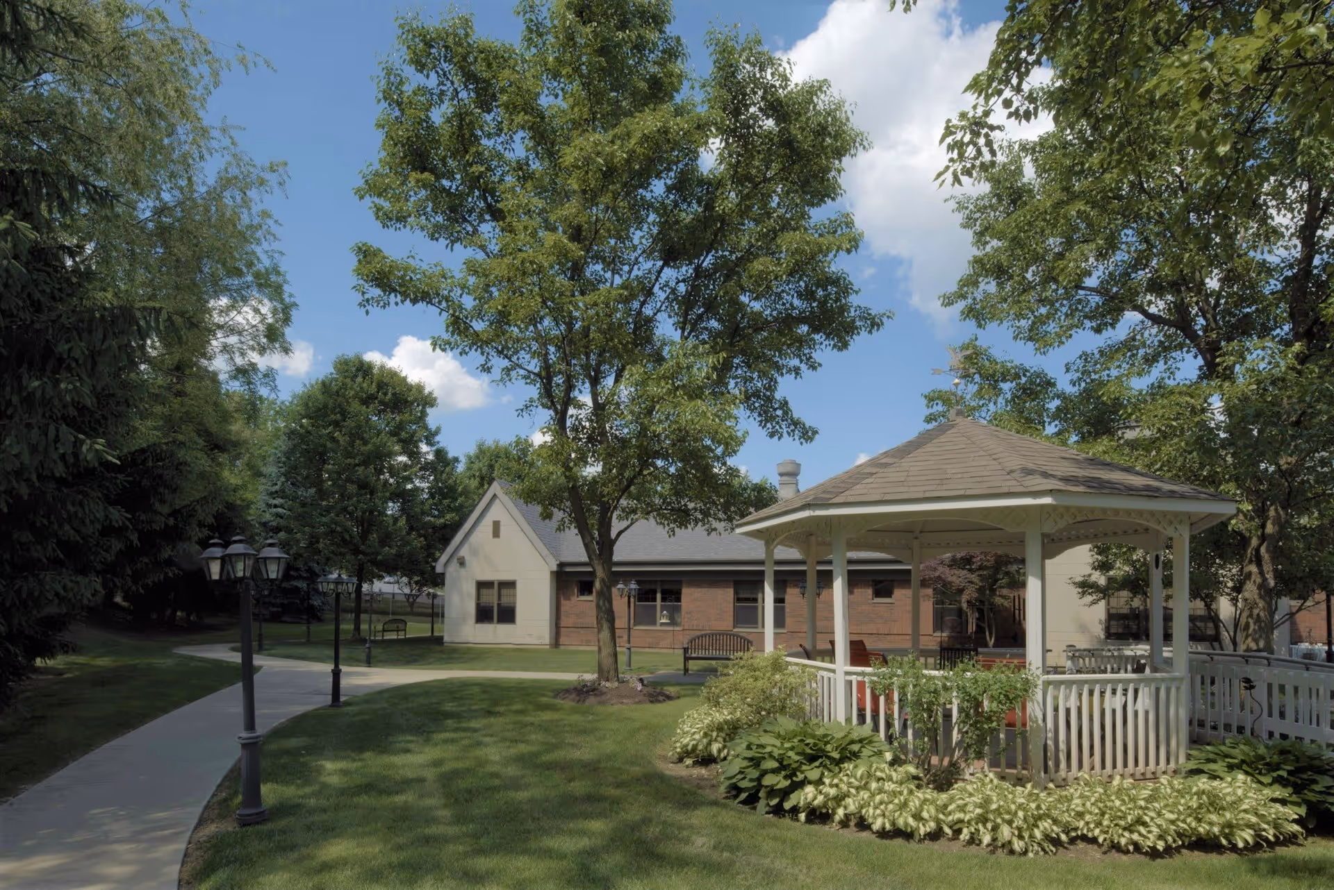 A landscaped outdoor courtyard with a white gazebo, walkway, trees, benches, and a low brick building in the background.