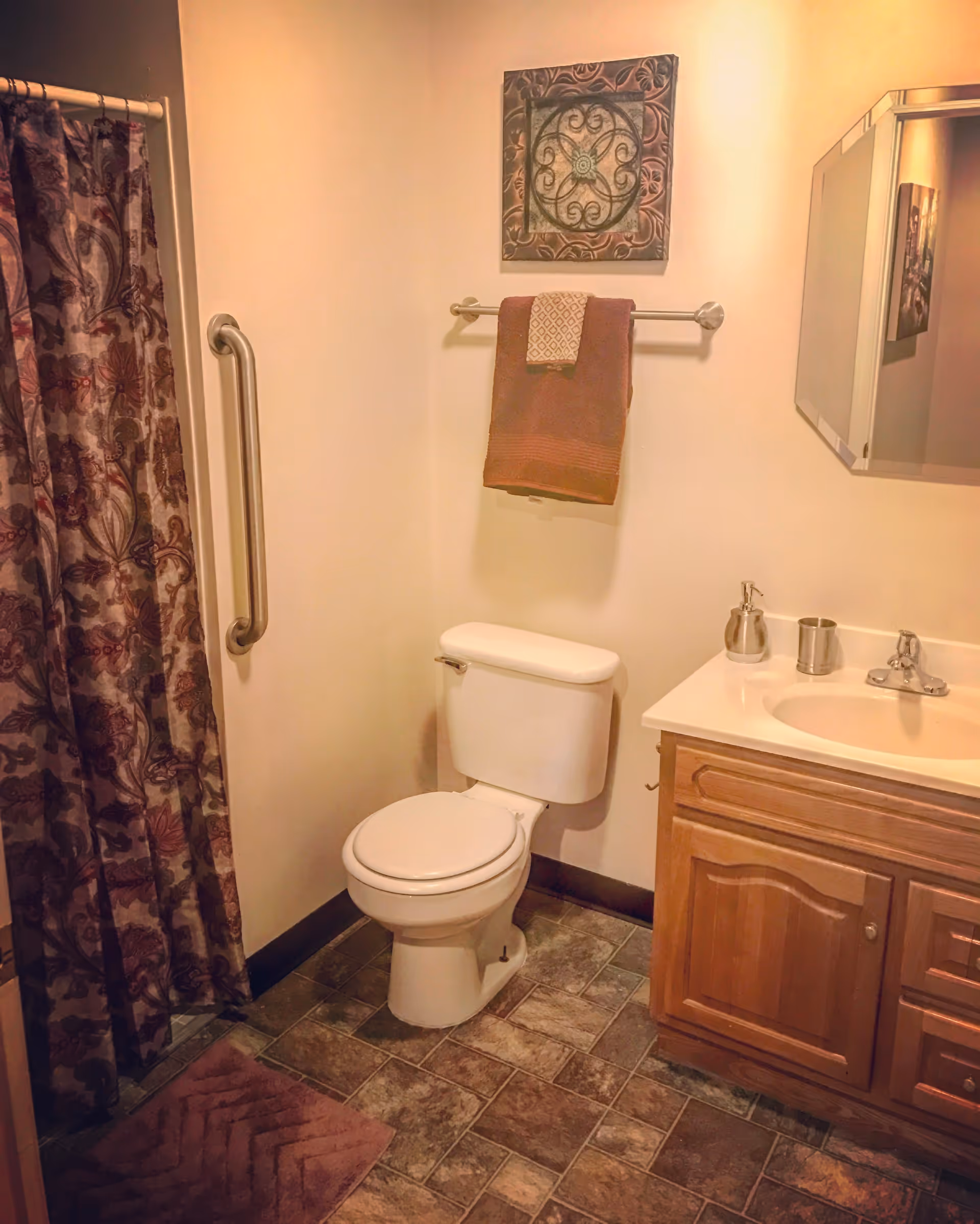 A bathroom with a white toilet, a wooden vanity with a sink, a mirror above the sink, a towel rack with two towels, a decorative wall hanging, a grab bar next to a shower with a patterned curtain, and a floor mat on tiled flooring.