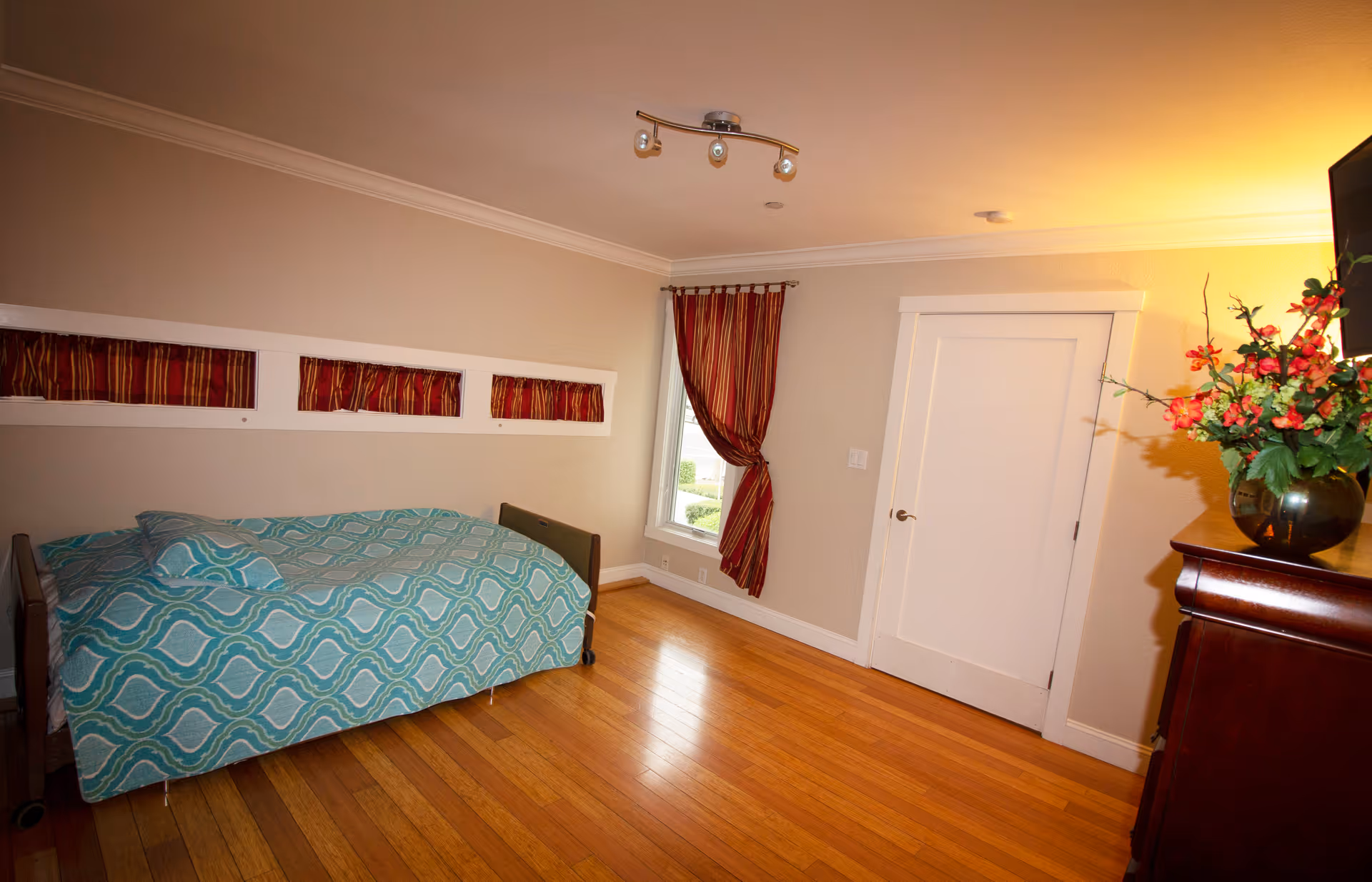 A bedroom with a single bed covered in a blue patterned bedspread, a window with red and gold striped curtains, a white door, a wooden dresser with a vase of red and orange flowers, and a wall-mounted TV.