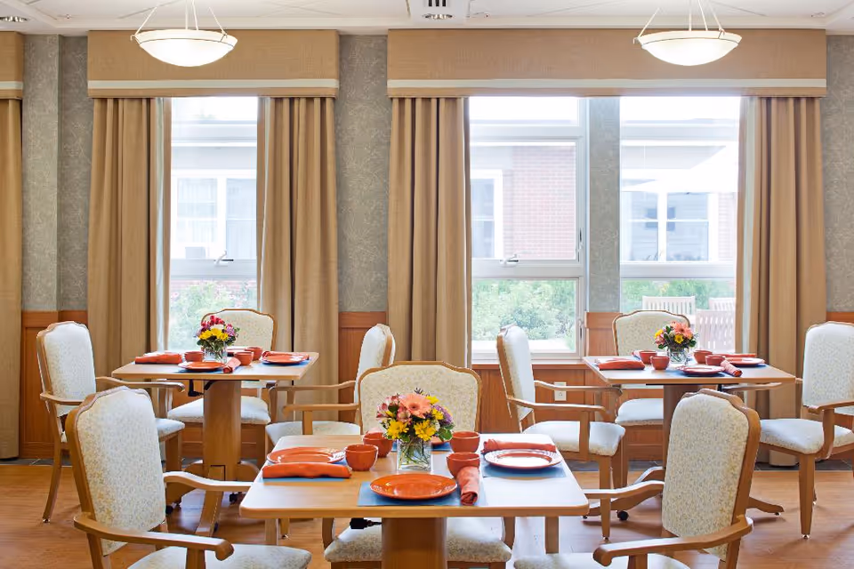 Dining room with wooden tables and upholstered chairs set with orange plates, bowls, napkins and floral centerpieces in front of large windows.