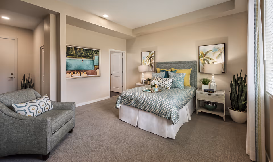 A well-lit bedroom with a bed dressed in patterned bedding and multiple pillows, two nightstands with lamps and decorative items, a comfortable armchair with a patterned pillow, potted plants, framed artwork on the walls, and a window with blinds and curtains.