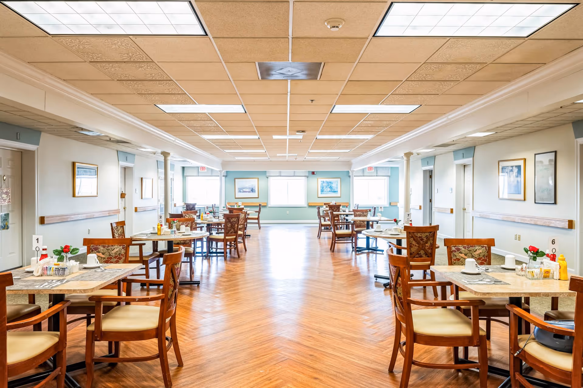 Bright and spacious dining room in a senior living facility with multiple wooden tables and chairs arranged neatly. Each table is set with placemats, cups, condiments, and small flower vases. The room has a wooden floor, light-colored walls with framed artwork, and large windows letting in natural light.