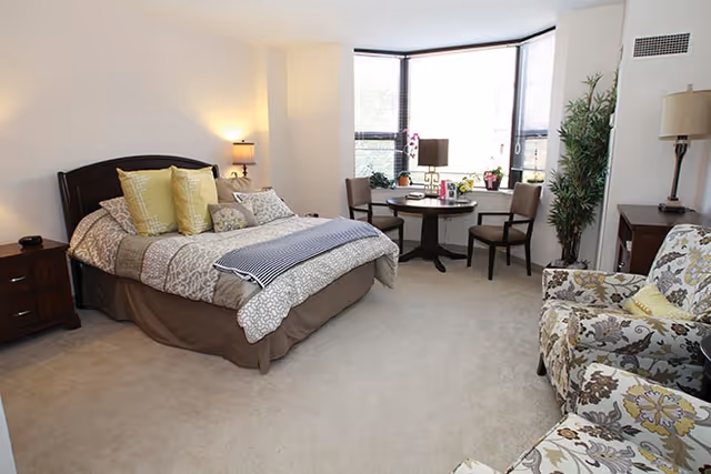 Spacious furnished bedroom featuring a bed, nightstands, a small table with chairs by a bay window, and two patterned armchairs.