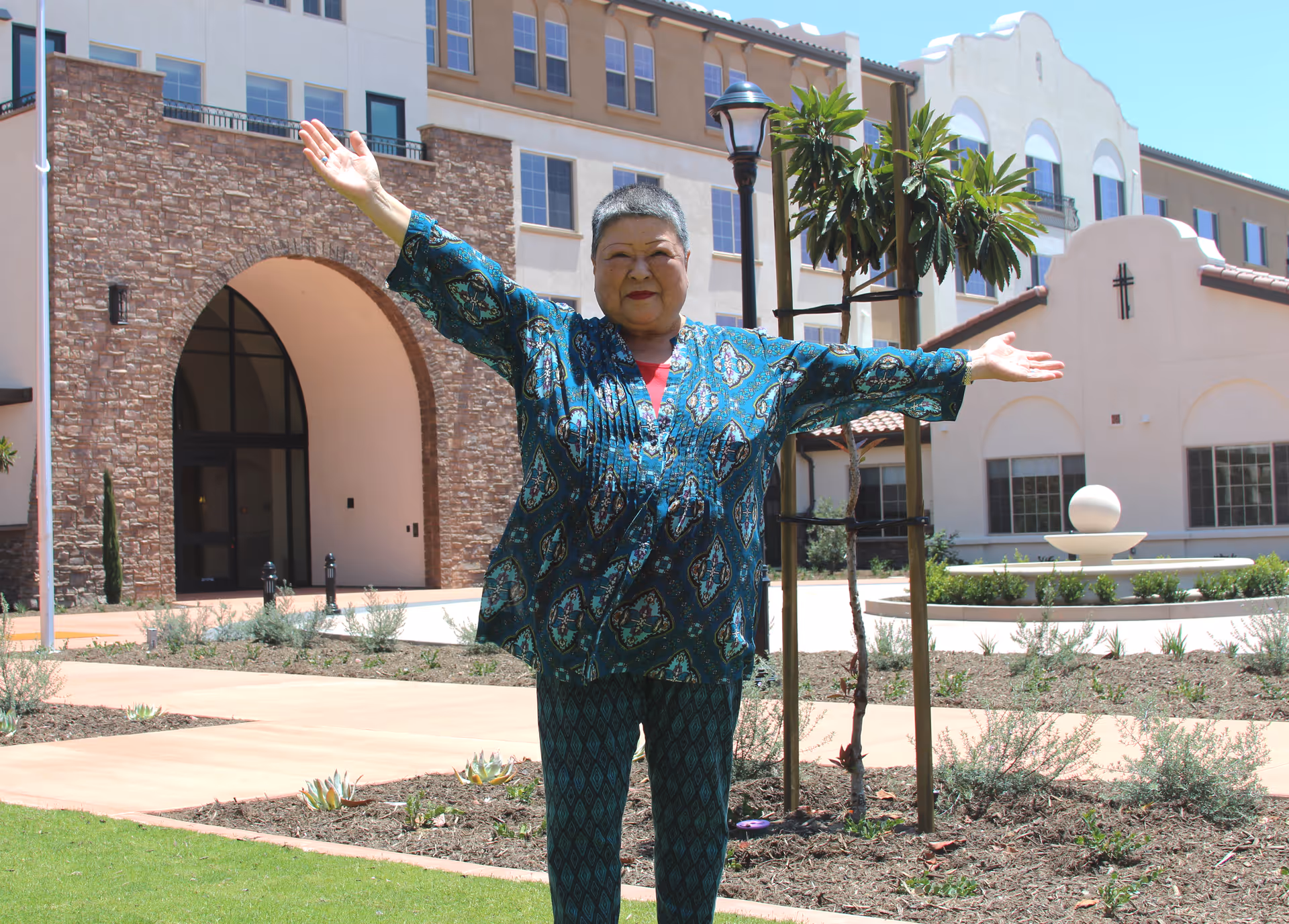 An elderly woman standing outside with her arms outstretched in front of a large building with stone and stucco exterior, featuring arched doorways and windows, and a small landscaped garden area.