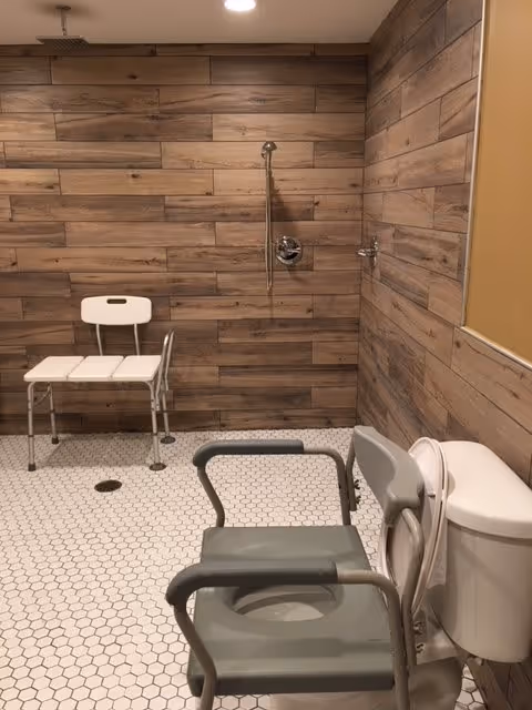 Accessible bathroom with wood-paneled walls and white hexagonal tile floor. The room features a white shower chair with backrest, a gray commode chair with armrests positioned next to a toilet, and a showerhead with a grab bar on the wall.