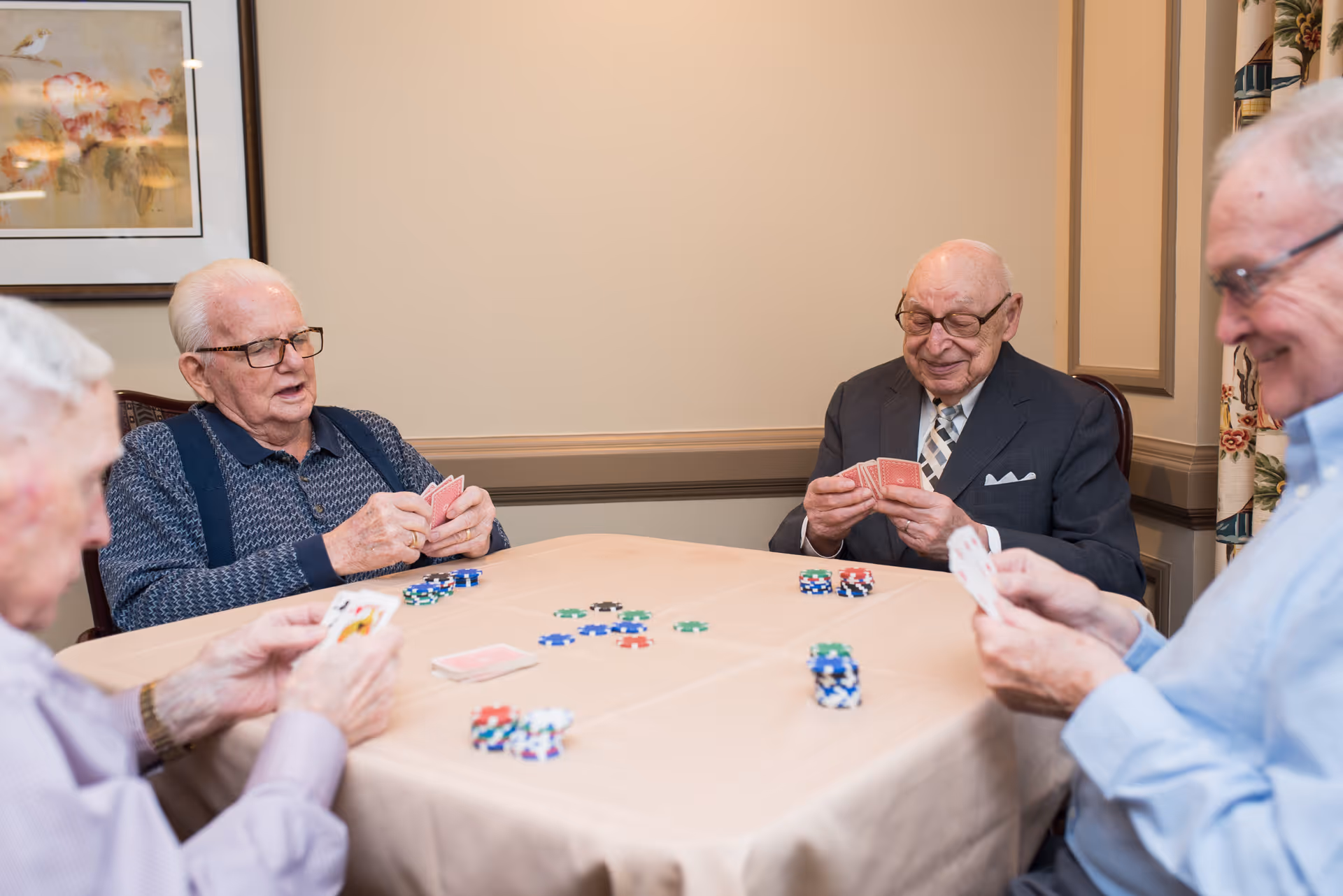 Four elderly men sitting around a table playing cards with poker chips scattered on the table in a well-lit room with beige walls and framed artwork.