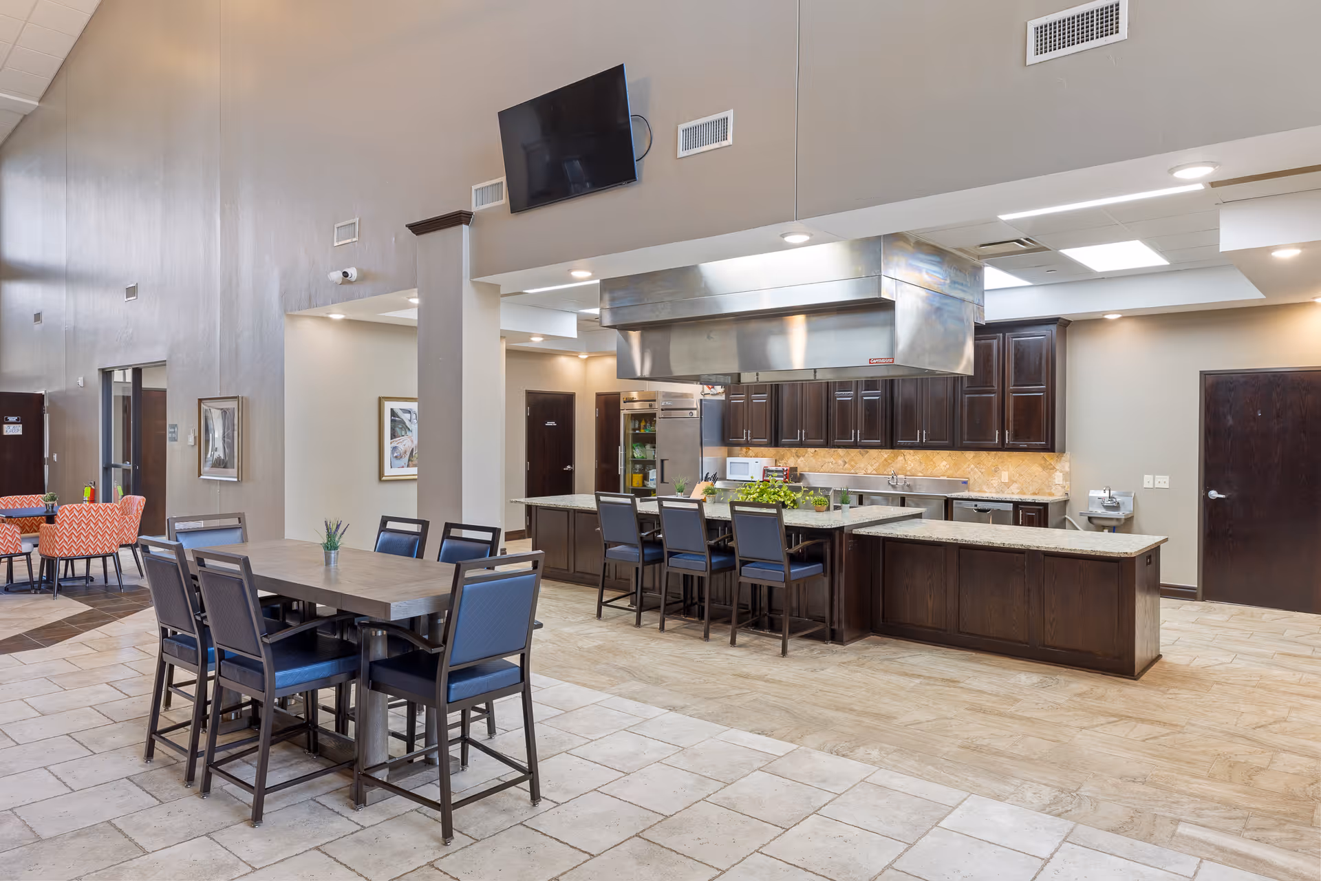 A spacious, modern kitchen and dining area in a senior living facility. The kitchen features dark wood cabinets, a large stainless steel range hood, granite countertops, and several appliances including a microwave and refrigerator. There are multiple seating areas with tables and chairs, including a high table with six chairs and a smaller table with orange patterned chairs in the background. The walls are painted beige, and the floor is tiled. A flat-screen TV is mounted high on the wall above the kitchen area.