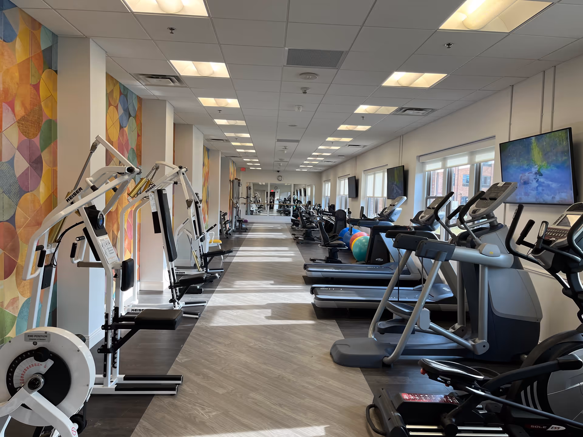 Long bright fitness room with treadmills, weight machines, exercise bikes, and colorful wall art.