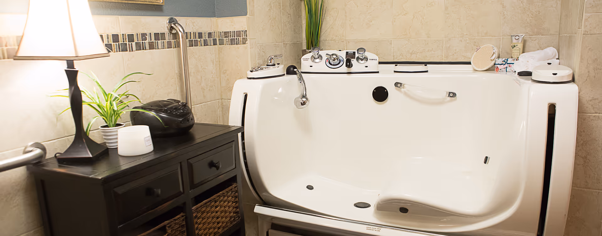 A bathroom featuring a white walk-in bathtub with control knobs and a safety handle. Next to the tub is a black wooden cabinet with drawers and wicker baskets, topped with a table lamp, a small potted plant, a candle, and a black radio. The walls are tiled with beige tiles and a decorative tile border.