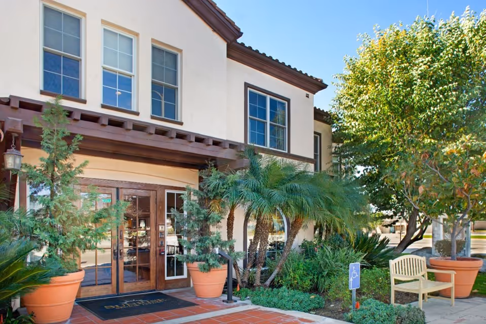 Exterior view of the entrance to a senior living facility with double wooden doors, large windows, and potted plants including small trees and palms. There is a bench and a handicap parking sign near the entrance, with a clear blue sky above.