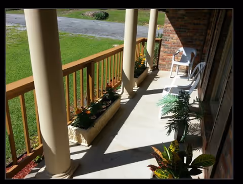 A sunny porch area with beige columns and a wooden railing. There are two white plastic chairs and a small white table on the porch. Several potted plants and flowers are placed along the edge of the porch and near the chairs. The porch overlooks a grassy lawn and a paved driveway.