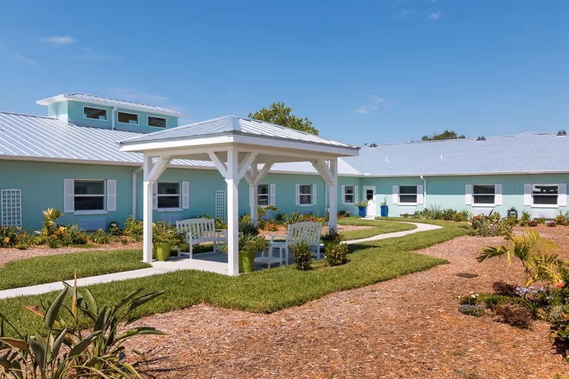 Outdoor garden area at Trinity Place Assisted Living featuring a white gazebo with benches and a table, surrounded by green grass, plants, and a light blue building with white trim under a clear blue sky.