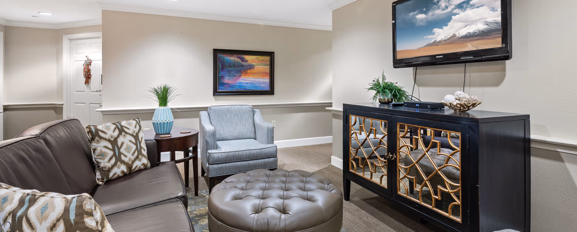 A cozy living room area with a dark brown leather sofa adorned with patterned cushions, a light blue armchair, and a round tufted ottoman in the center. A wooden side table with a blue vase holding green plants is next to the armchair. On the wall, there is a colorful landscape painting and a flat-screen TV mounted above a decorative black cabinet with mirrored doors. The room has beige walls and carpeted flooring.
