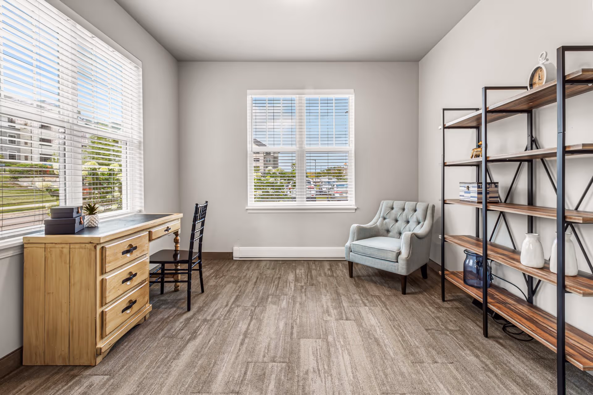 A bright room with large windows featuring white blinds, a wooden desk with three drawers and a black chair, a light blue upholstered armchair, and a tall black metal shelving unit with wooden shelves holding decorative items.