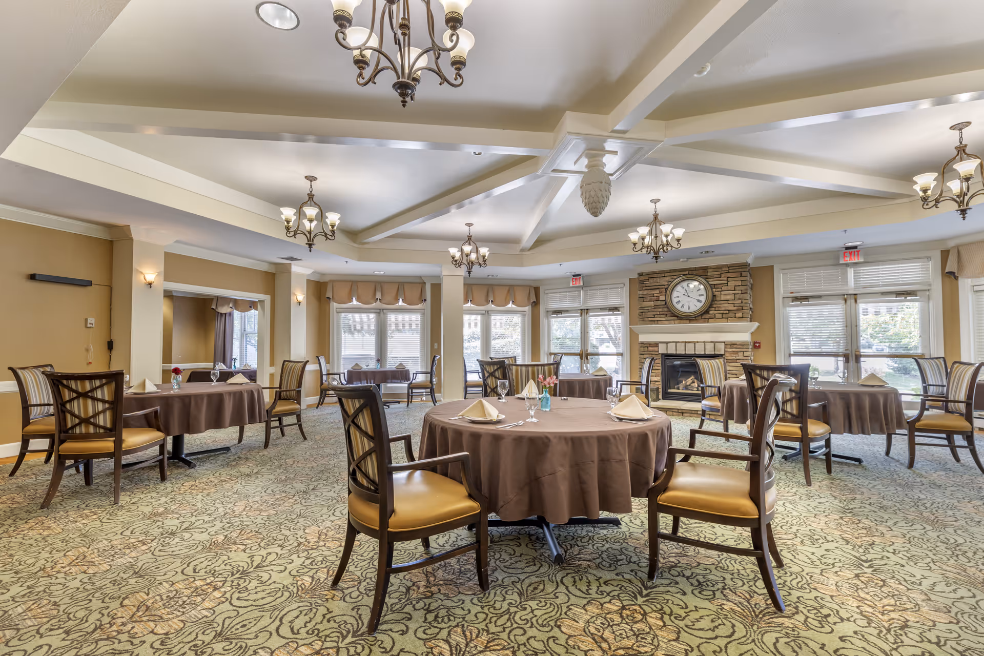 A spacious dining room with round tables covered in brown tablecloths, each set with napkins, glasses, and silverware. The room features patterned carpet, multiple chandeliers hanging from a coffered ceiling, large windows with beige valances, and a stone fireplace with a clock above it.
