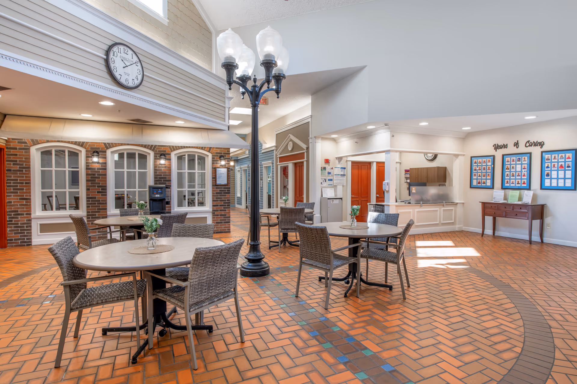 A bright and spacious common area with round tables and wicker chairs arranged on a patterned brick floor. A tall black streetlamp-style light fixture stands in the center. The walls feature a brick facade with windows and a clock above, and a bulletin board labeled 'Years of Caring' is visible on the right. The ceiling is high with recessed lighting, and there are doors and a reception area in the background.