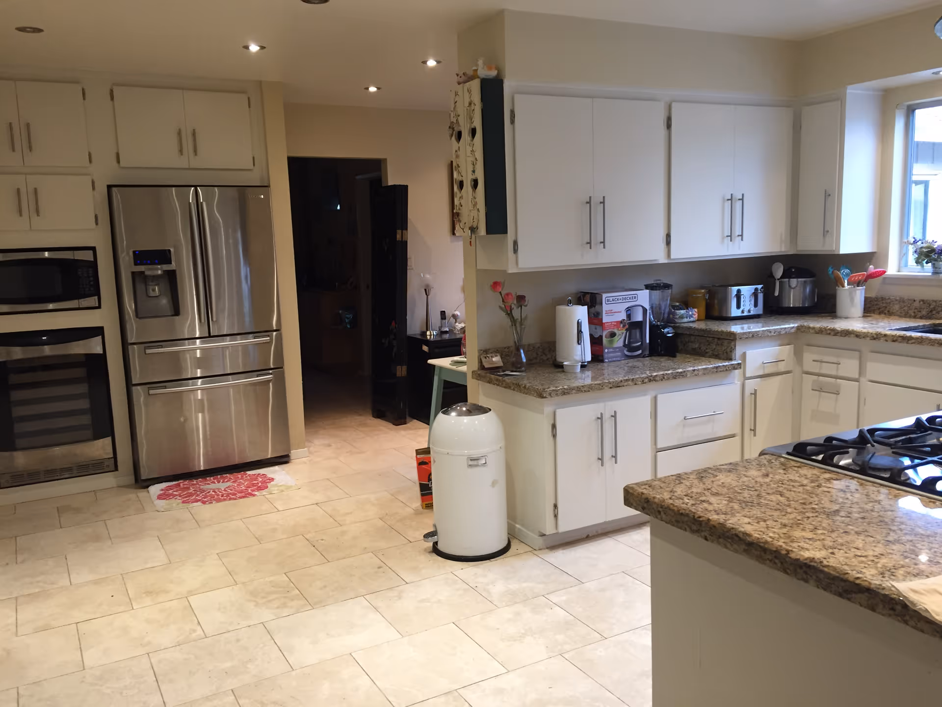 A modern kitchen with white cabinets, granite countertops, and stainless steel appliances including a refrigerator, oven, and microwave. There is a gas stove on the right countertop, a white trash can on the floor, and various kitchen appliances such as a toaster, coffee maker, and blender on the counters. A window above the sink lets in natural light.