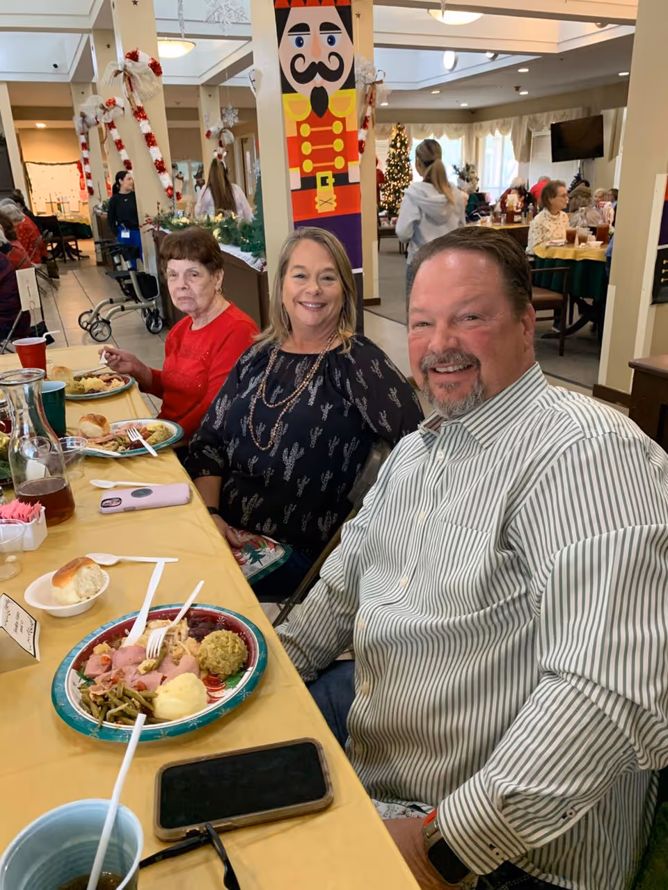 Three people sit at a decorated dining table in a senior living facility with plates of holiday food in front of them.