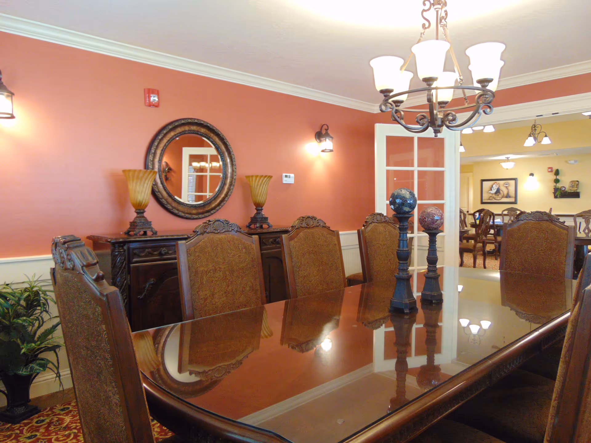 Formal dining room with a long polished table, upholstered chairs, chandelier, and a decorative sideboard against a red wall.