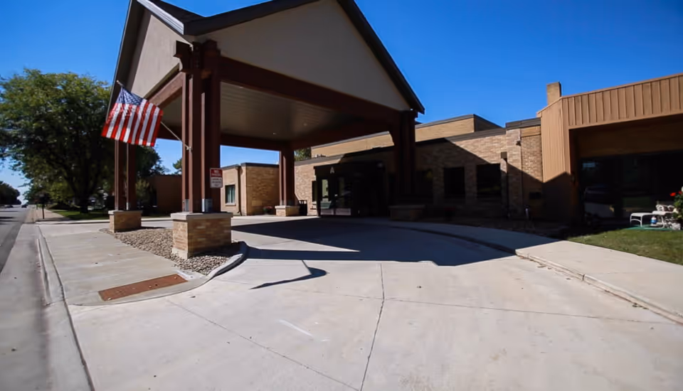 Entrance to a senior living community building with a covered drop-off area supported by wooden beams. An American flag is displayed on the left side near the sidewalk. The building is made of brick and has large windows. The sky is clear and blue.