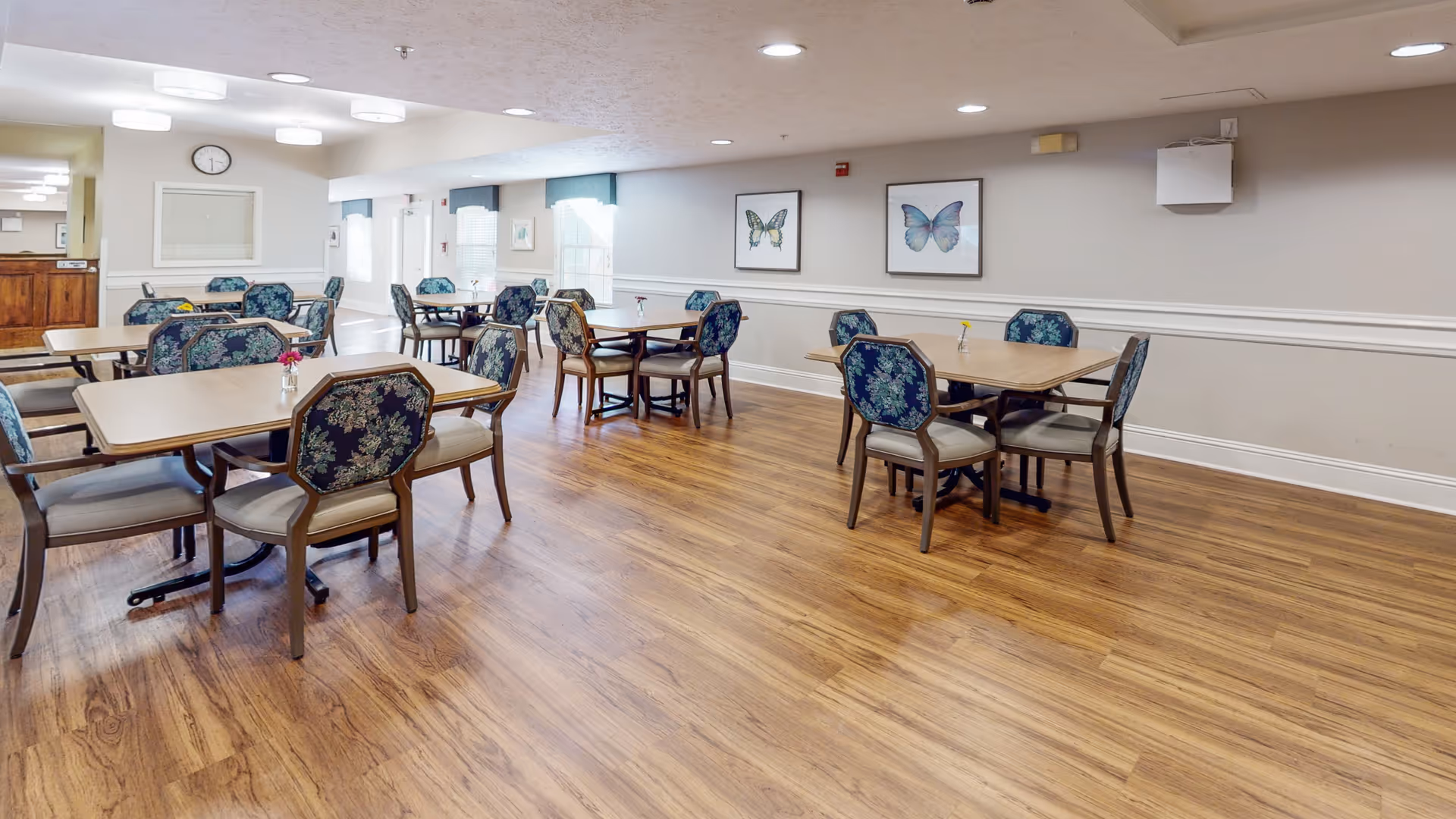 A spacious dining room with wooden floors and several tables surrounded by chairs with floral upholstery. The walls are light-colored with two framed butterfly artworks hanging. The room is well-lit with ceiling lights and natural light coming through windows with white curtains.