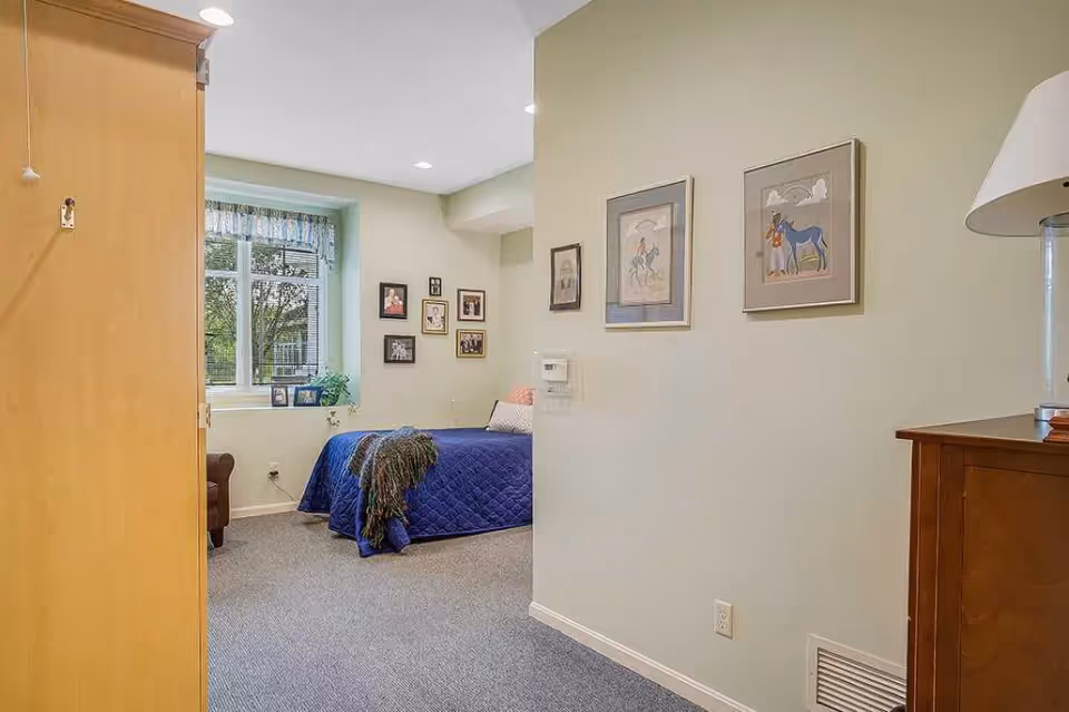 A cozy bedroom in a senior living facility with a bed covered in a blue quilt, a window with a valance letting in natural light, several framed pictures on the walls, a wooden dresser with a lamp, and a small armchair near the window.