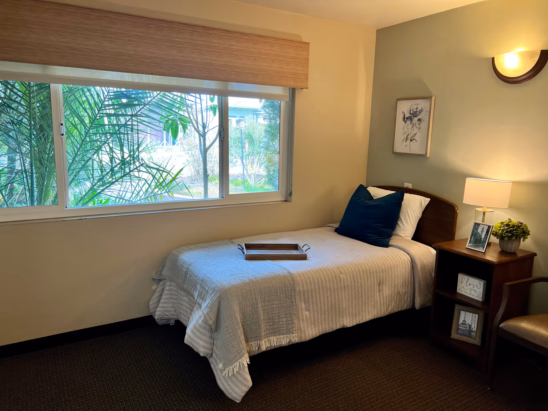 A small, cozy bedroom with a single bed covered in a light gray striped blanket and a dark blue pillow. A wooden tray rests on the bed. Next to the bed is a wooden nightstand with a lamp, a small plant, framed photos, and a decorative sign. A large window with a beige valance shows green plants and trees outside. The walls are painted in light neutral tones with a framed floral artwork above the bed.