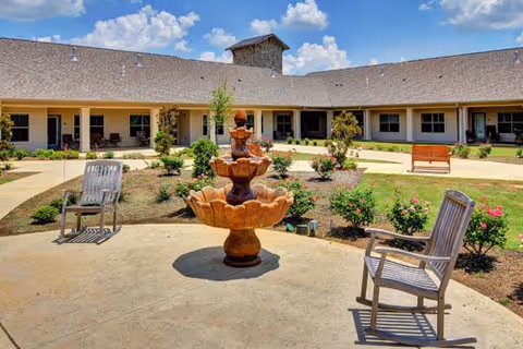 Outdoor courtyard area of a senior living facility with a central multi-tiered fountain surrounded by garden beds with flowers and shrubs. There are wooden chairs and benches placed around the courtyard, and the building with multiple windows and a shingled roof encloses the space under a partly cloudy blue sky.