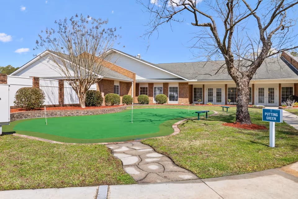 Front exterior of a single-story senior living building with a small putting green, trees, and a paved walkway.