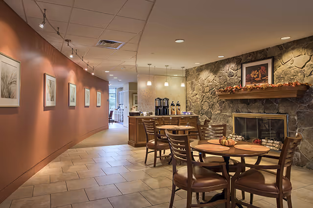 Cozy communal dining area with round tables and chairs, a stone fireplace, and a coffee station along a curved wall.